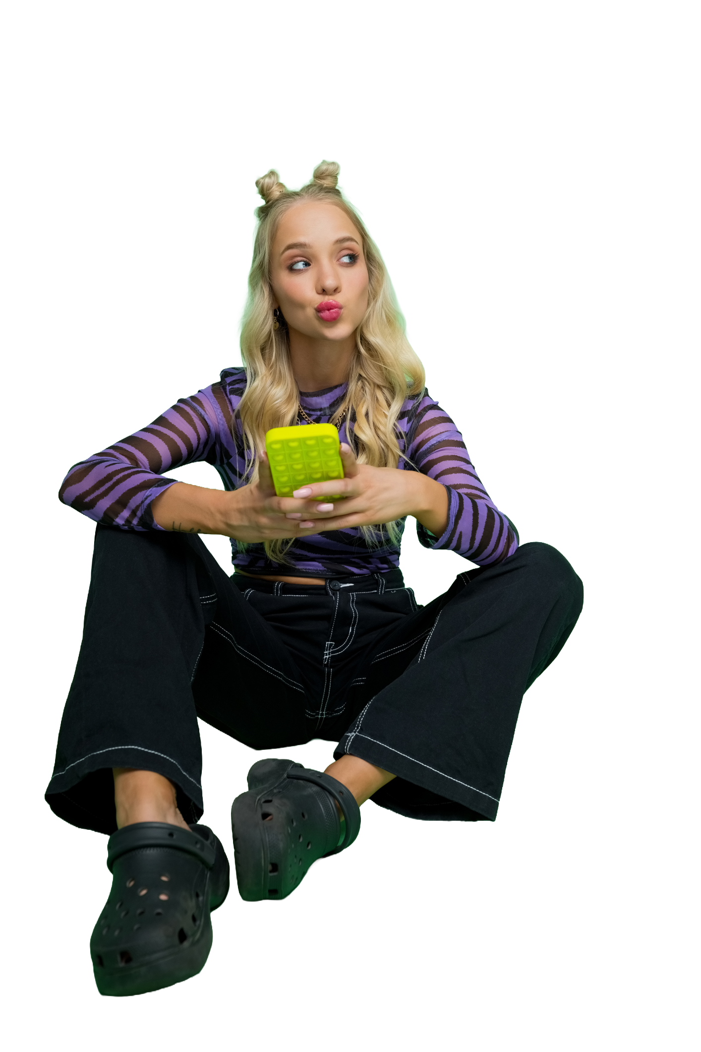 Full length shot of thoughtful young blonde woman wearing purple animal pattern blouse and black pants holding mobile phone in hand, sitting on floor and using mobile phone. Studio shot, green background.