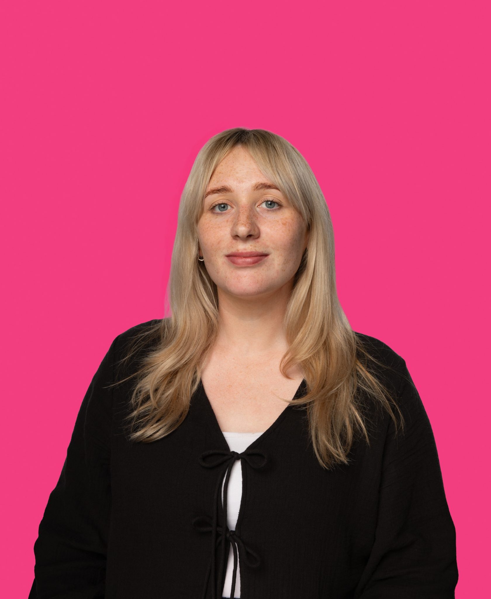 Woman with long blonde hair wearing a black top, posed against a vibrant pink background.