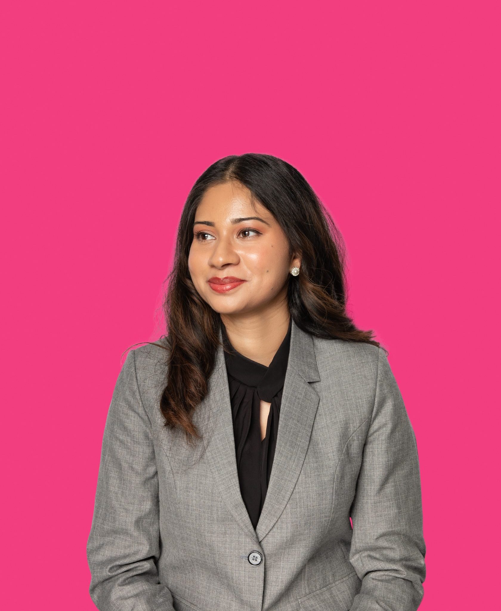 Confident woman with long brown hair wearing a grey blazer and black top, posed against a vibrant pink background.