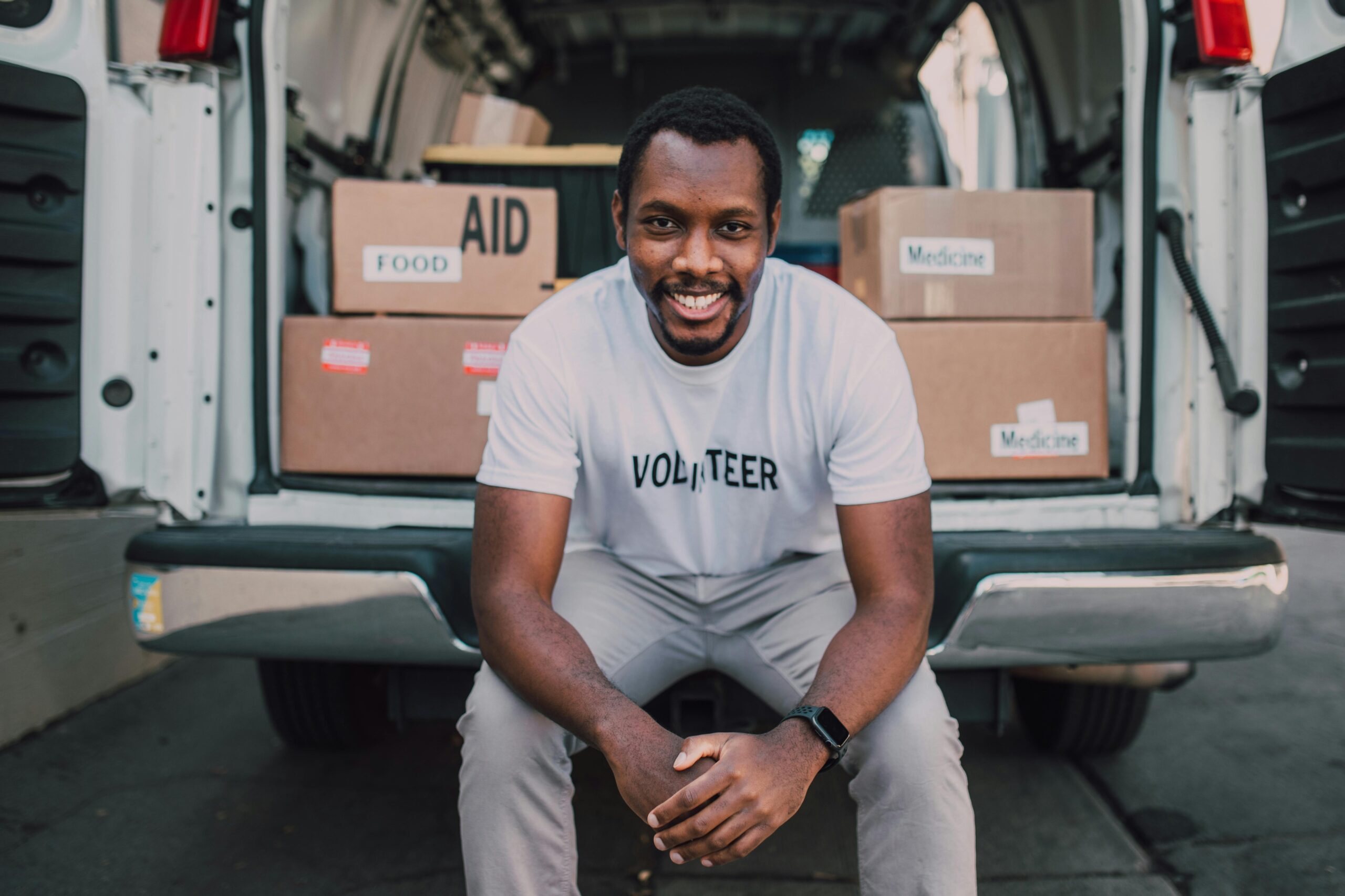 Black Man, sitting on back of donation van wearing volunteer shirt smiling