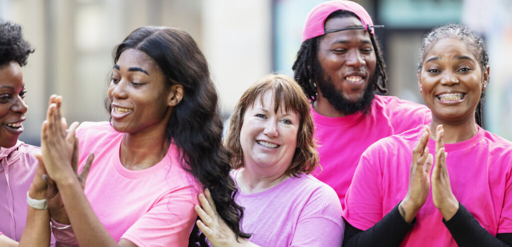 A multiracial group of friends wearing pink, attending a breast cancer awareness event. A mid adult African-American man is standing with the four women.